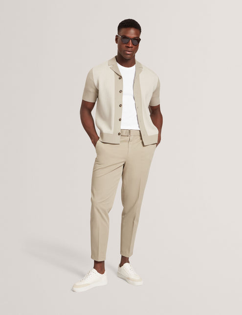 A male model wearing a short sleeve knitted shirt in beige color with a revere collar and button-through design, paired with light beige trousers and sunglasses.