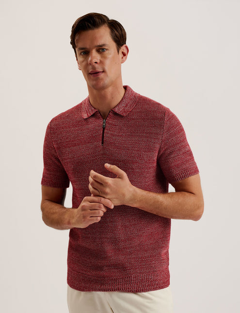 A male model wearing a red short sleeve polo shirt with a classic collar and a zip neck, paired with white trousers and white shoes.