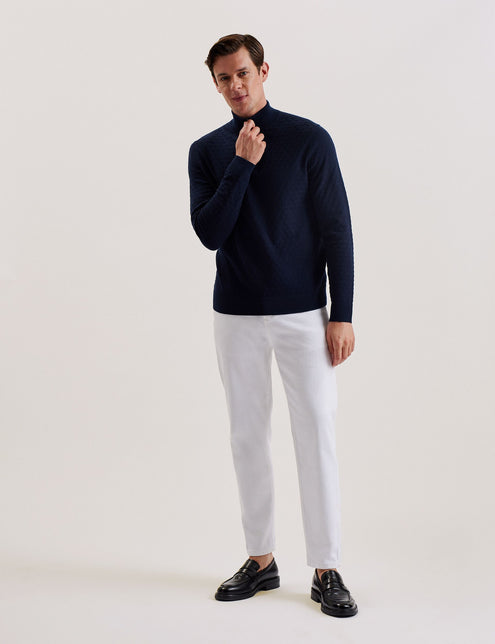 A man wearing a navy long sleeve wool blend jumper with a funnel neck and half zip placket, paired with white trousers.